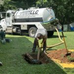 Worker bending over to connect hoses to a trench in a grassy backyard, with a large vacuum truck in the background preparing sewer line work; a yellow tarp lines the excavation.